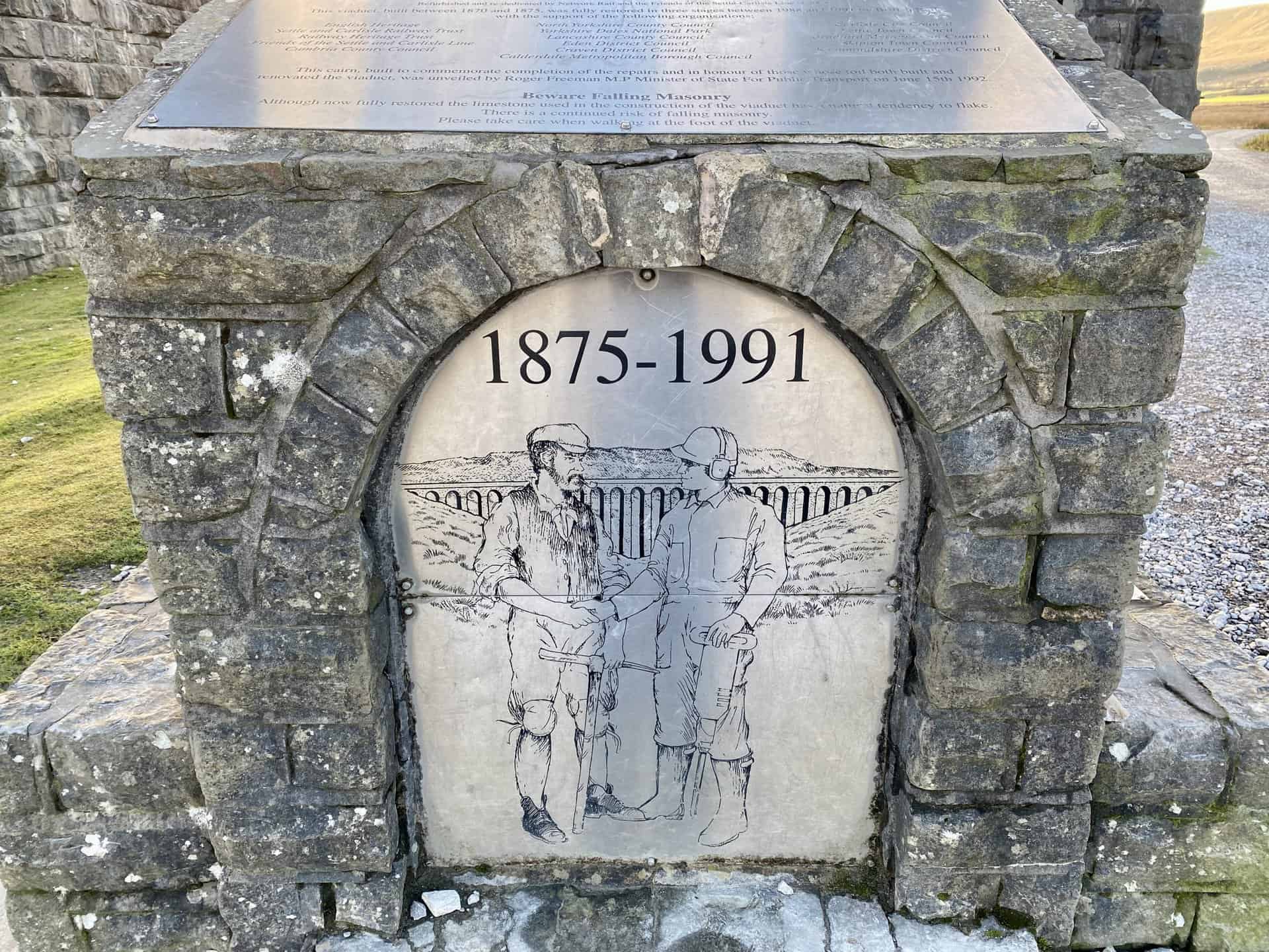 The silver plaque attached to one side of the stone statue near the foot of the viaduct.