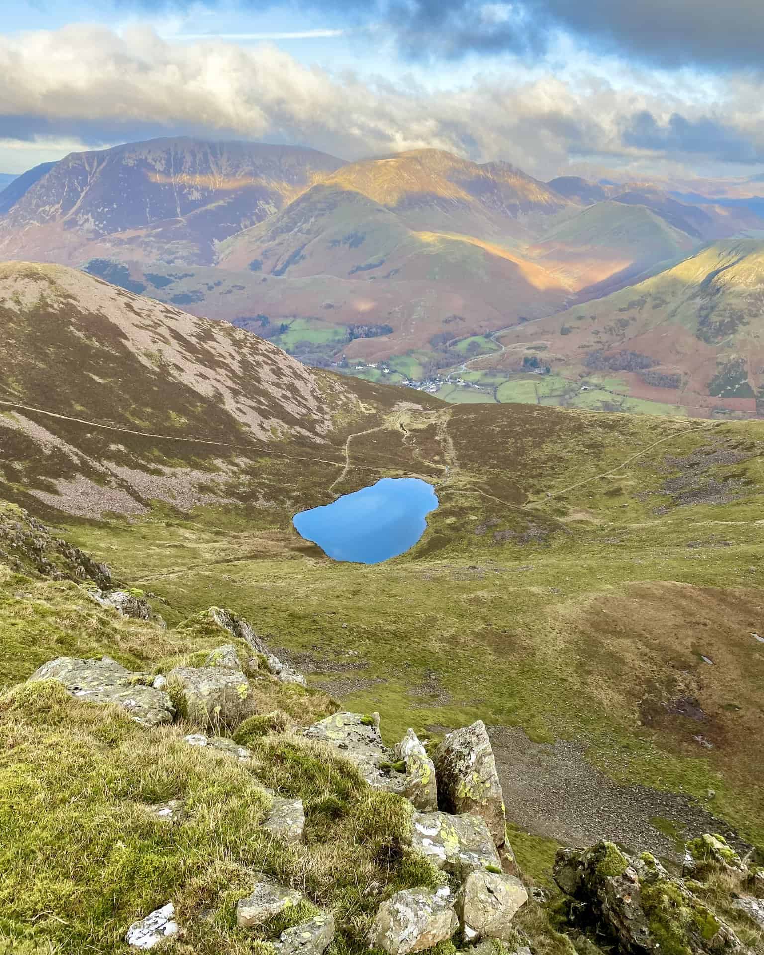 Spectacular vistas from above Chapel Crags. Bleaberry Tarn down below, and the village of Buttermere in the valley. Stretching out behind Buttermere is the mountain range comprising Whiteless Pike, Wandhope, Crag Hill and Sail. The largely brown-coloured mountain on the left-hand side is Grasmoor.