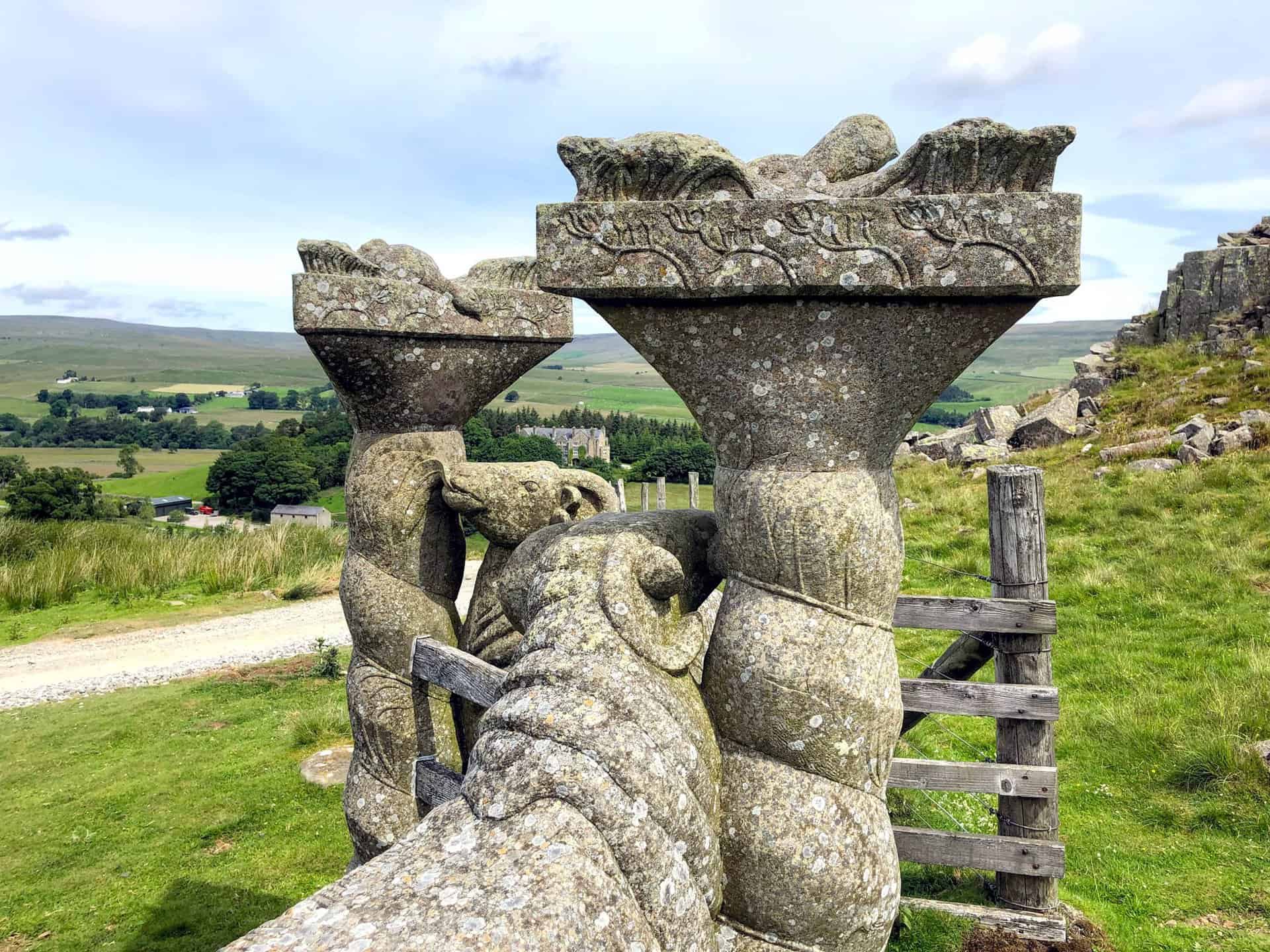 Intricately crafted stone sheep flank a stile near Holwick Scars, blending art with the natural landscape.