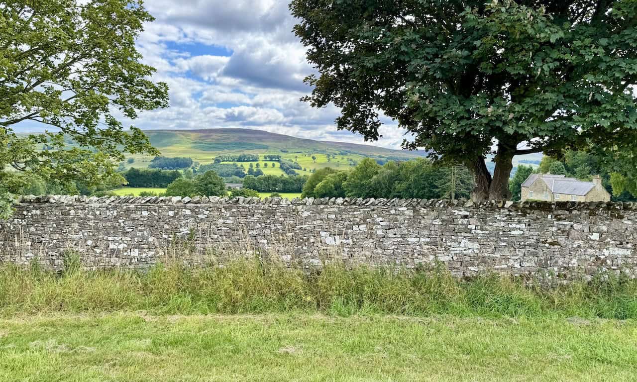 Rolling farmland and scattered trees stretch out beyond the wall near Braithwaite Moor.
