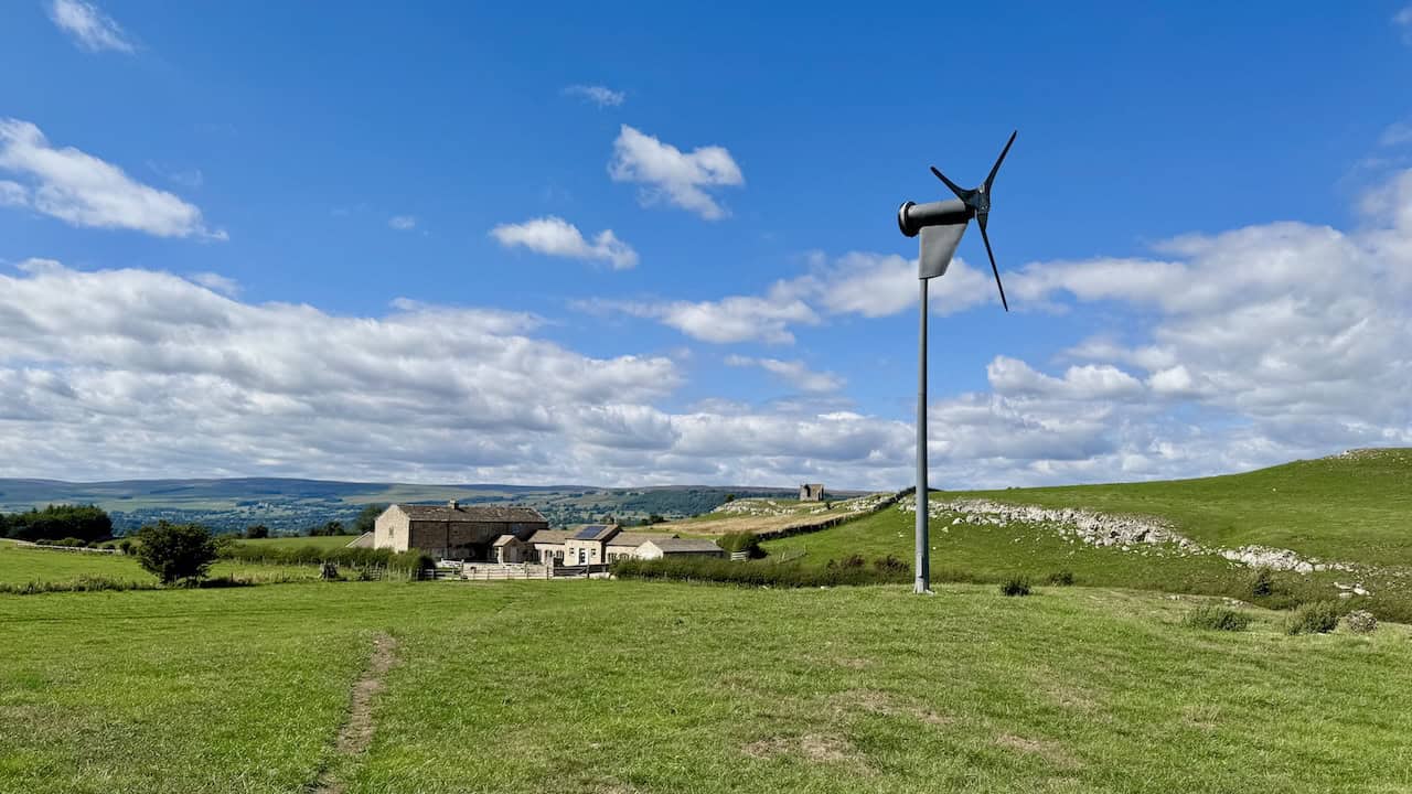 A wind turbine spins near well-maintained farm buildings with limestone outcrops.
