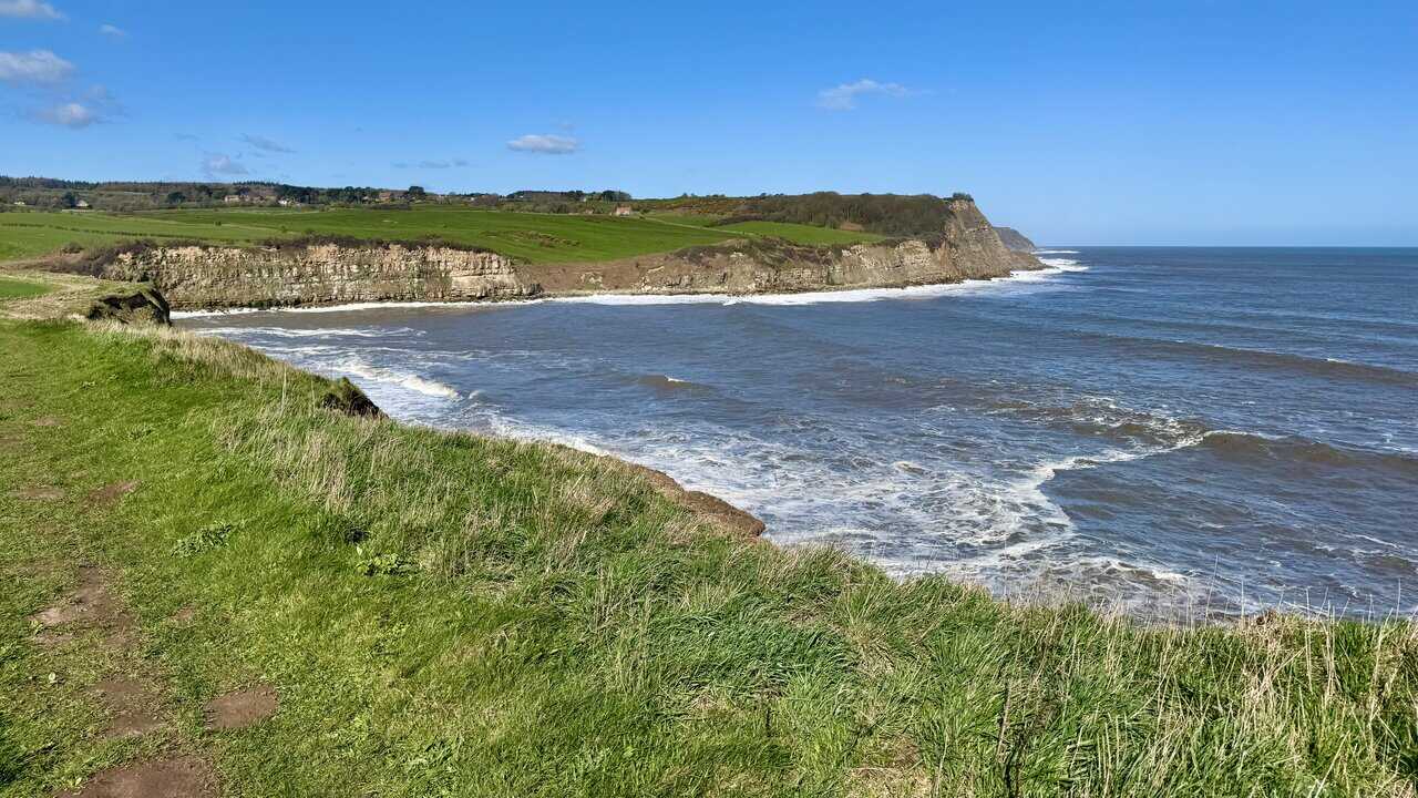 Curving coastline approaching Cloughton Wyke, with layered cliffs, rocky shoreline and green farmland rising inland.