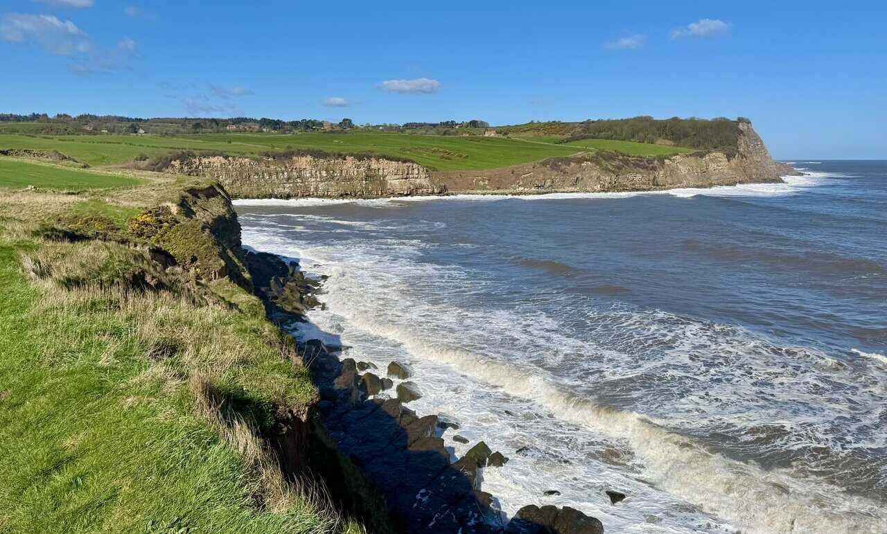 Wide bay view towards Cloughton Wyke, with horizontal rock bands beneath green fields and waves breaking white at the foot of the headland.