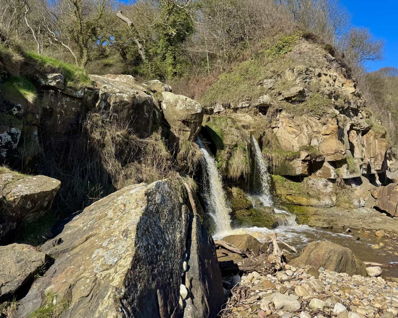 Double waterfall at Hayburn Wyke dropping over a rocky ledge onto the beach, framed by mossy outcrops, boulders and Jurassic cliffs.