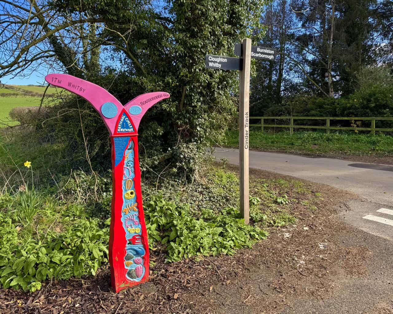 Cinder Track crossroads at Station Lane in Cloughton, with a brightly painted National Cycle Network milepost showing distances to Whitby and Scarborough.