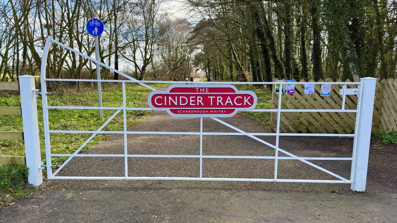 White gates at Cloughton Station with Cinder Track nameplate-style signs, opening onto a smooth, flat and accessible section of trail.