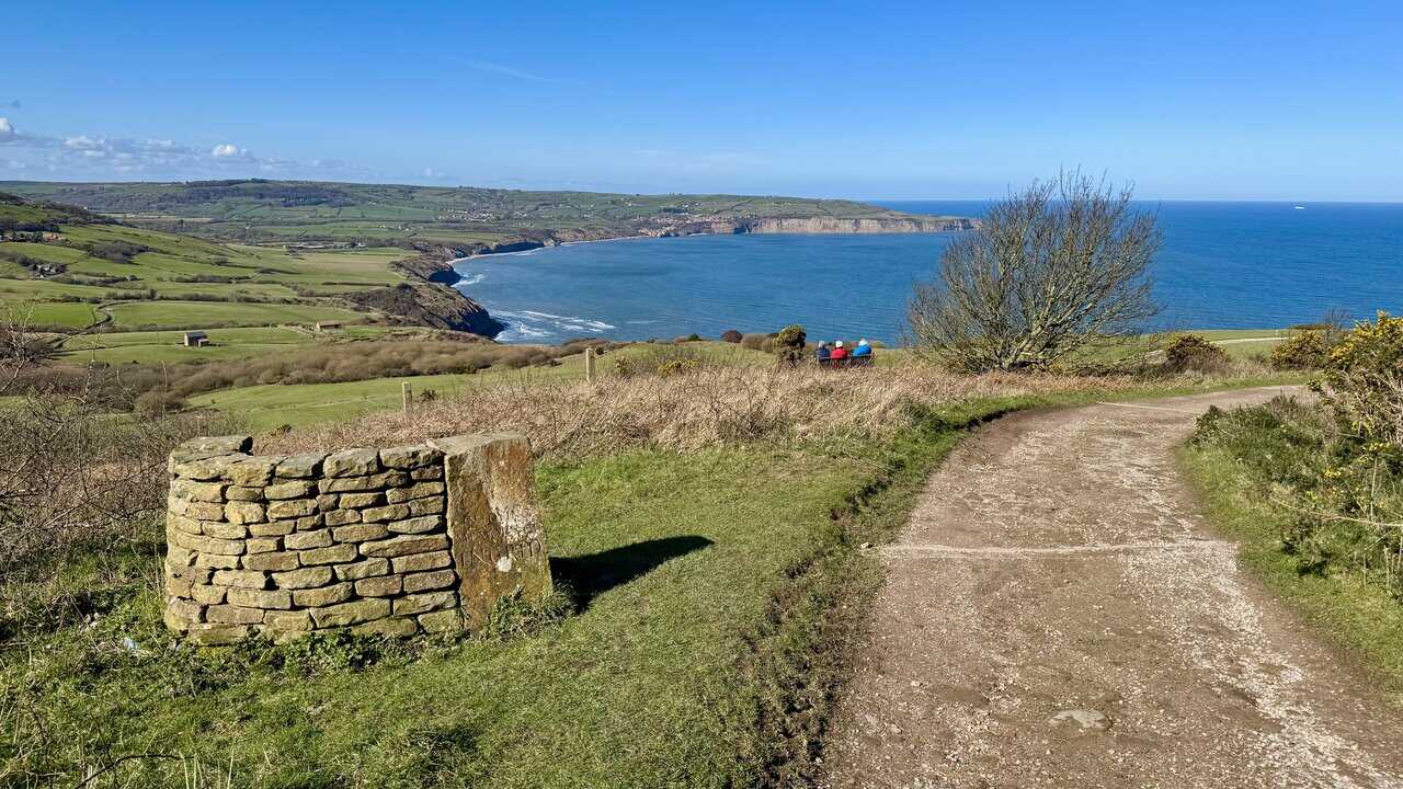 View from the path below Raven Hall Hotel across Robin Hood’s Bay, with visitors seated on a bench.