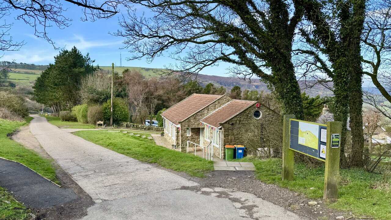 Concrete track leading downhill past Ravenscar Visitor Centre, with an information board beside the clifftop path and rolling green hills beyond.