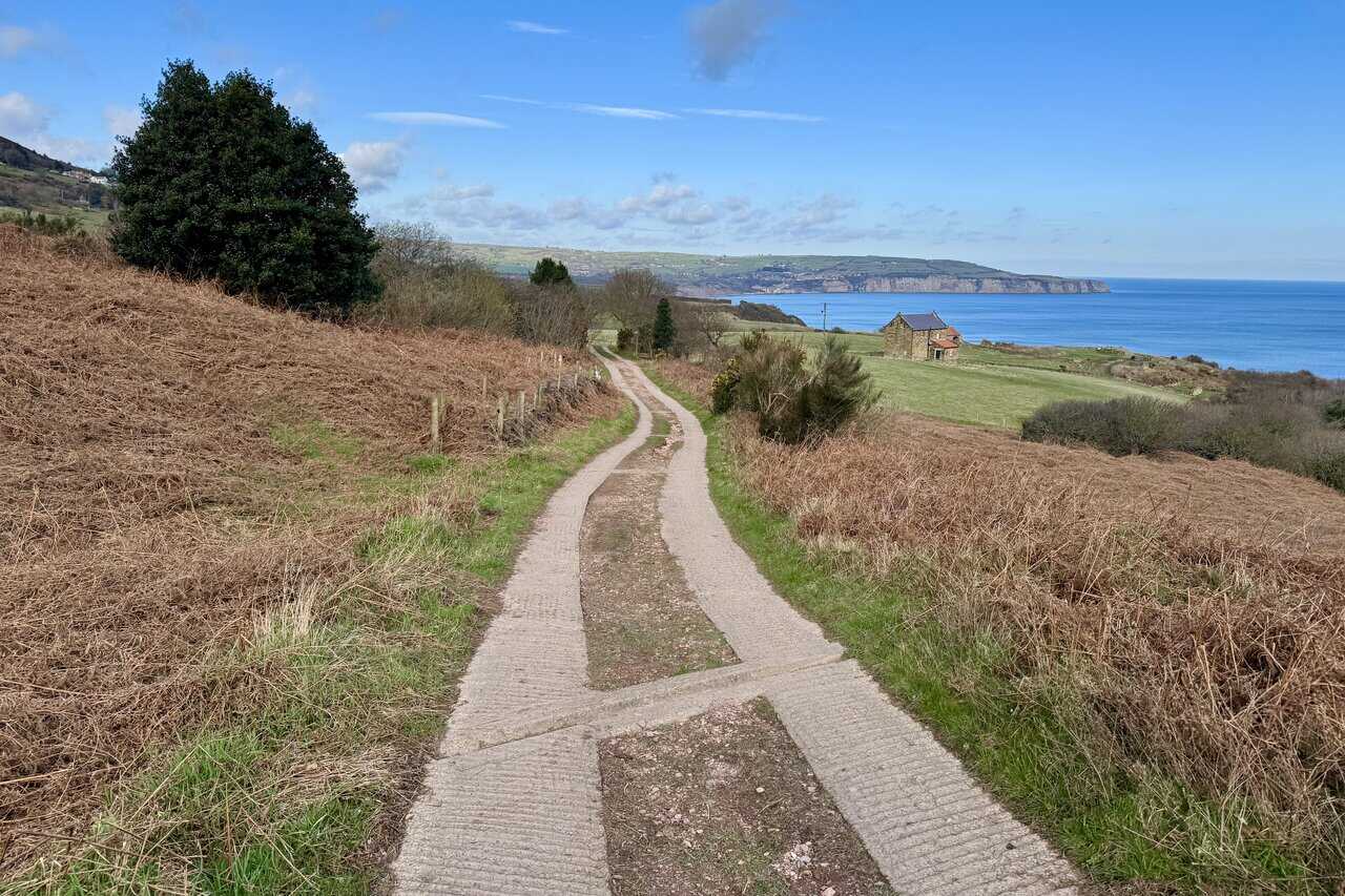 Concrete track curving towards Low Peak farmhouse near the cliff edge, with wide views north across Robin Hood’s Bay and its pale cliffs.