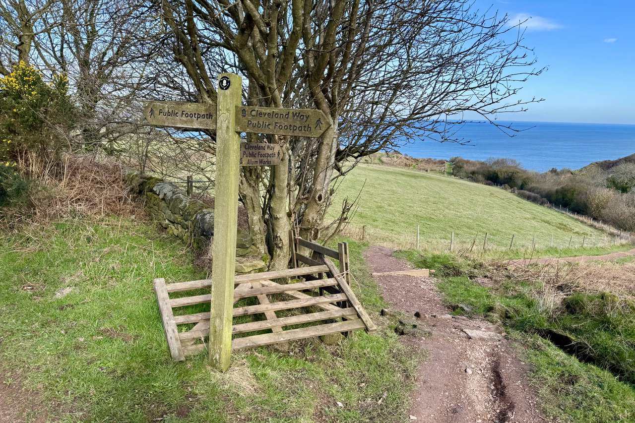 Wooden signpost on the Cleveland Way near Low Peak, beside a broken gate and a green field, pointing towards Peak Alum Works and the sea.