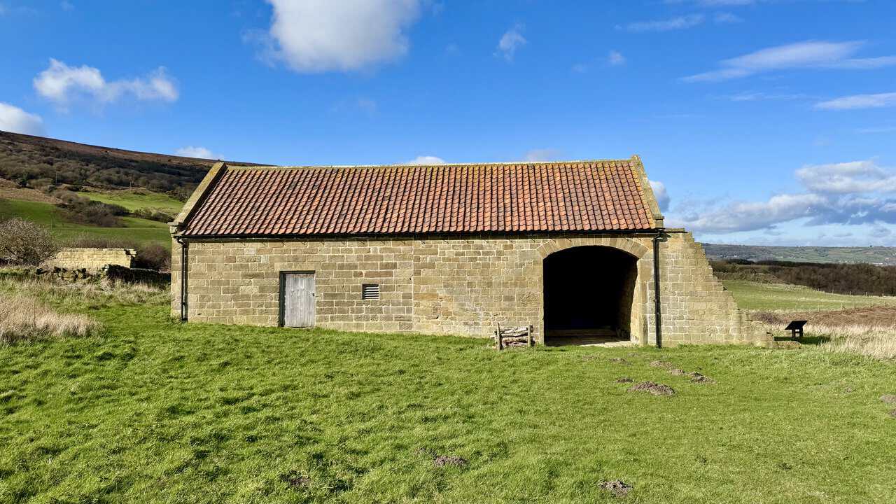 Stone barn with a red pantile roof and wide arched opening, standing in open green fields beneath the slopes of Stoupe Brow.