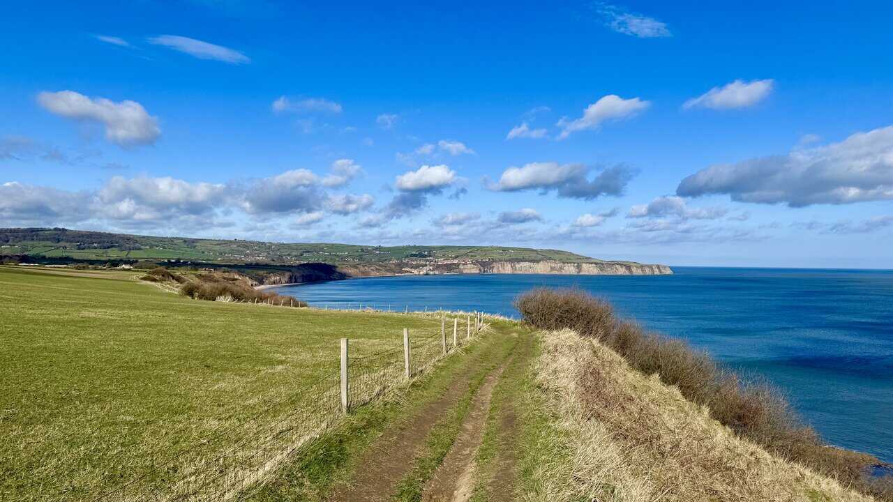 Narrow clifftop path between a green field and the sea, with sweeping views across Robin Hood’s Bay towards the distant village. A highlight of the Ravenscar walk.