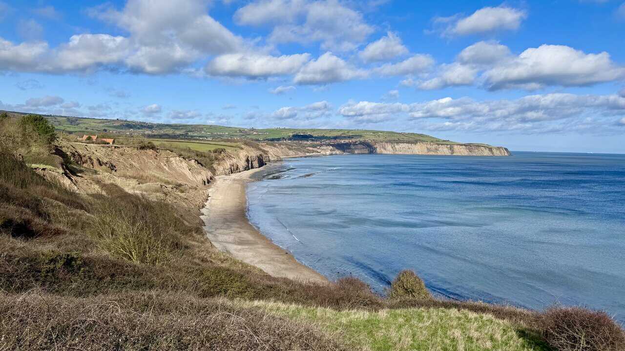 View north from the pillbox towards Stoupe Beck Sands, with golden sand, shallow clear water and layered cliffs backed by green fields.