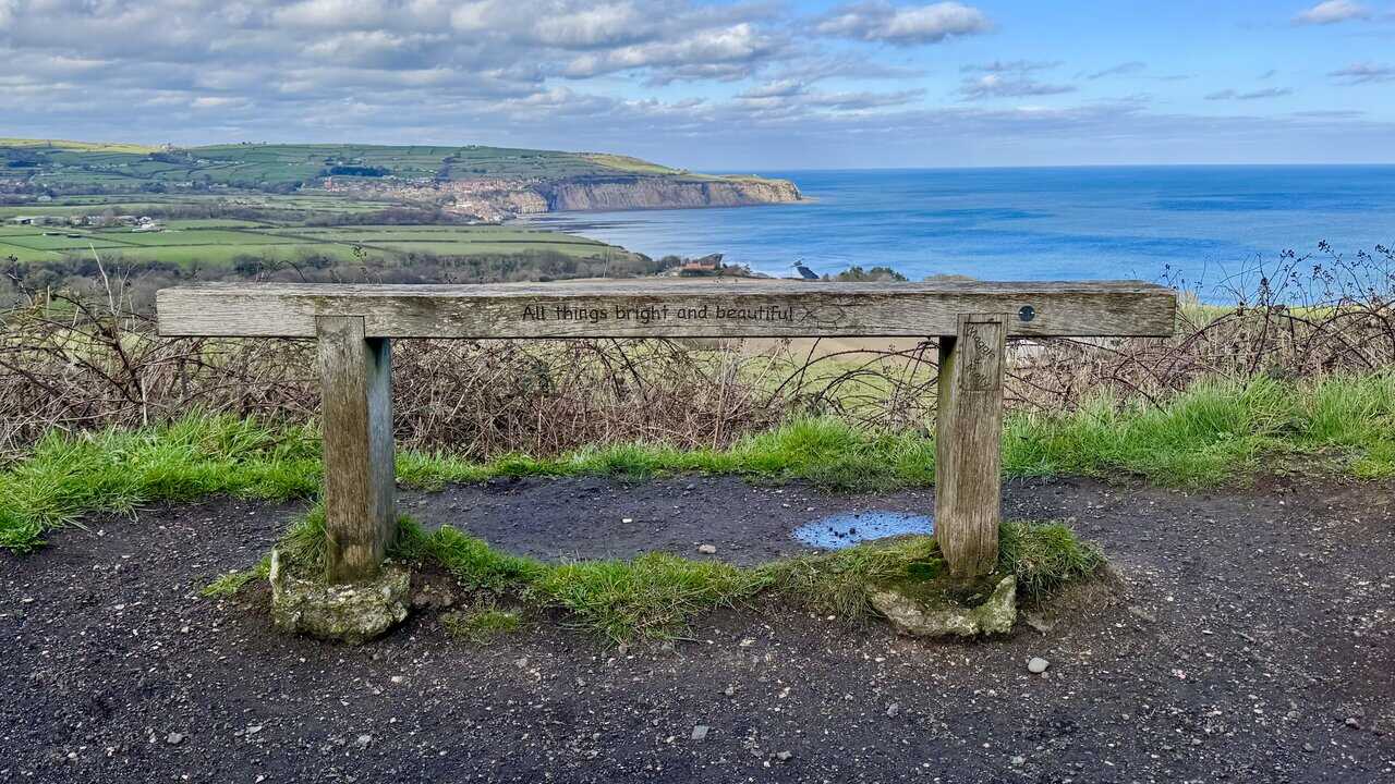 Wooden bench carved with the words ‘All things bright and beautiful’, overlooking Robin Hood’s Bay and the wide sweep of the coast.
