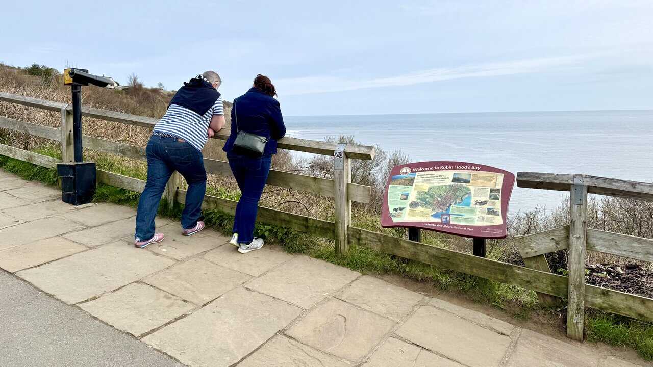 Visitors by the fence, telescope and information board at the top viewpoint on the Robin Hood's Bay walk.