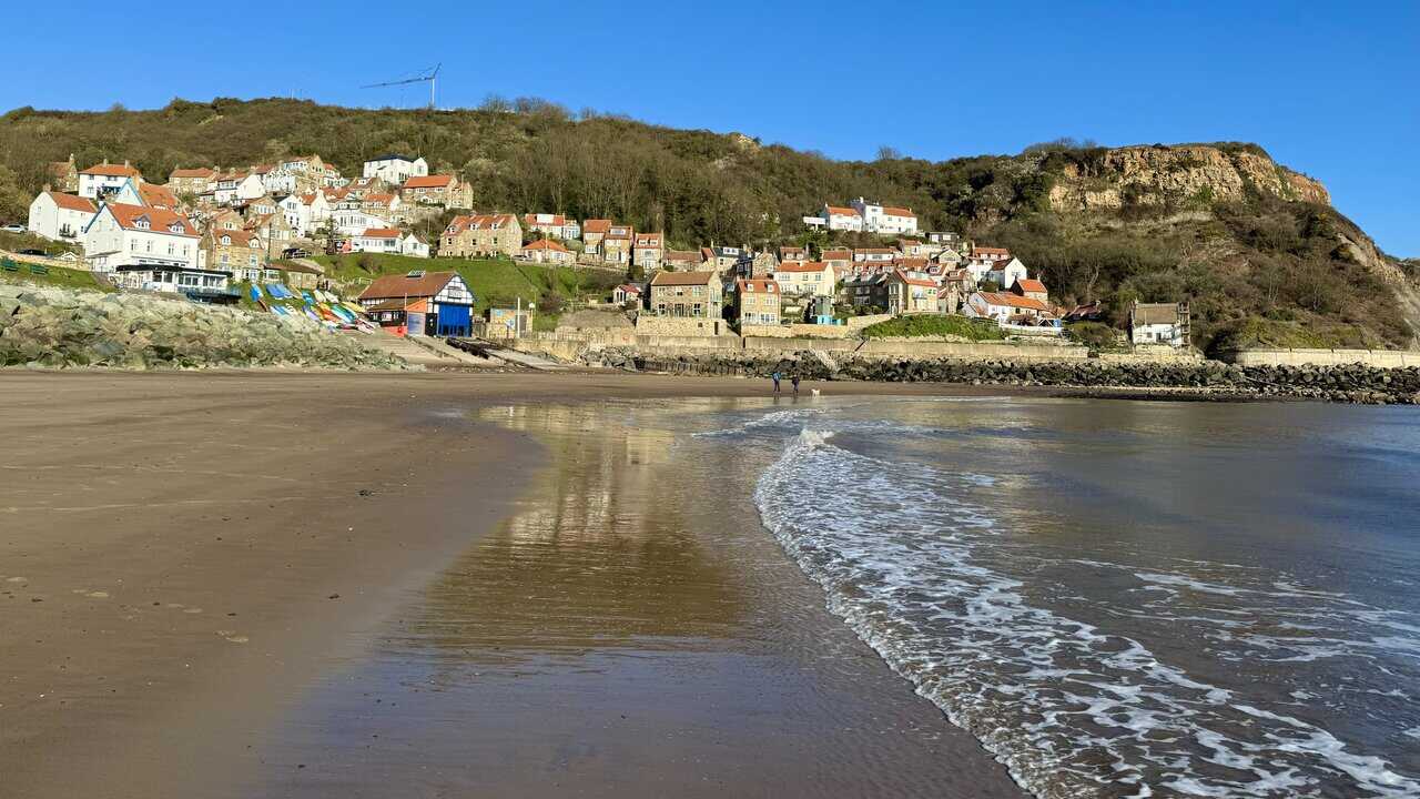 Wet sand on Runswick Bay beach reflecting the village above, with a broad curved bay, rocky edges and a sheltered shoreline at low tide.