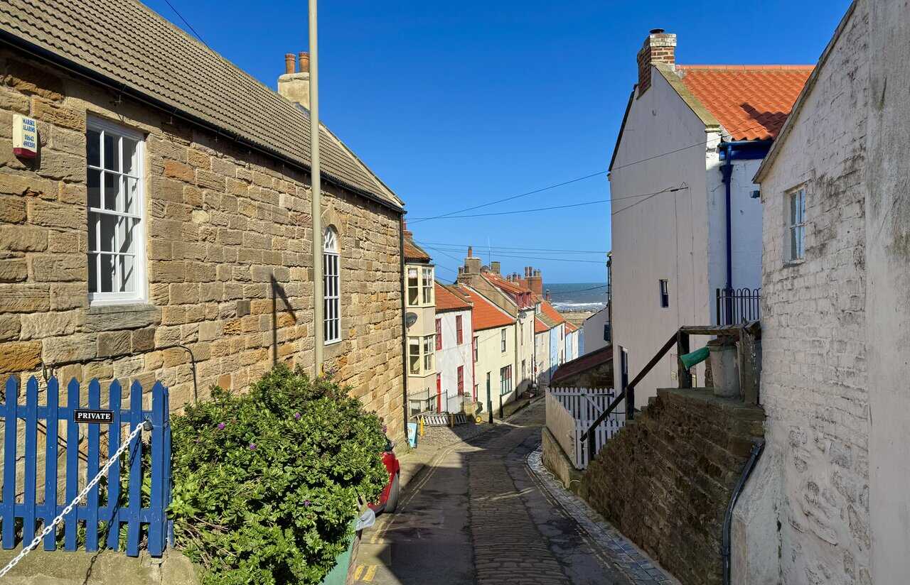 Steep cobbled lane on the Runswick Bay walk descending into Staithes between sandstone and white-rendered cottages, with glimpses of the sea ahead.