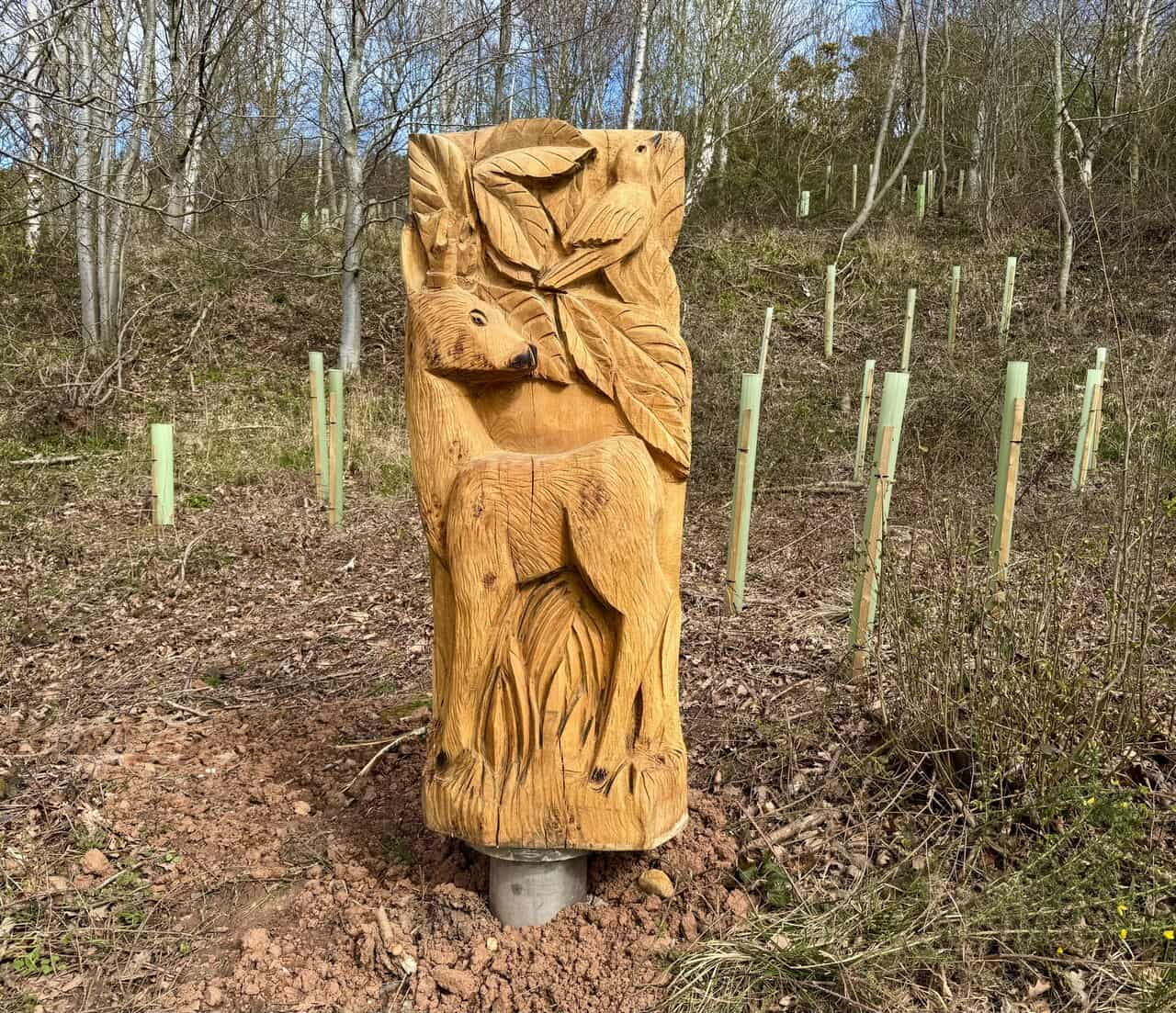 Tall wooden panel carving of a deer beneath broad leaves, with a small bird above, displayed beside the woodland path.