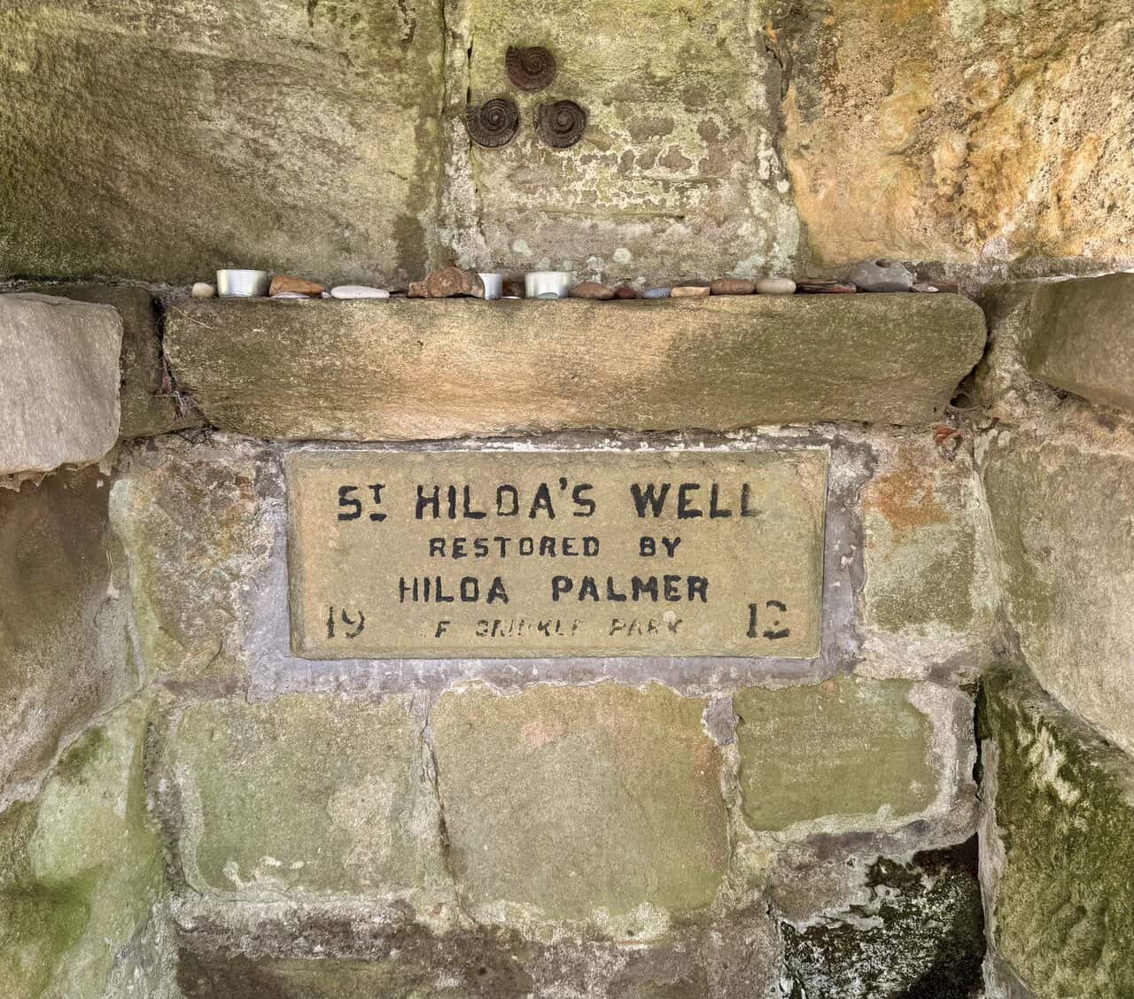 Offerings of stones, shells and candles at St Hilda’s Well, with ammonite fossils set into the stonework above the plaque.