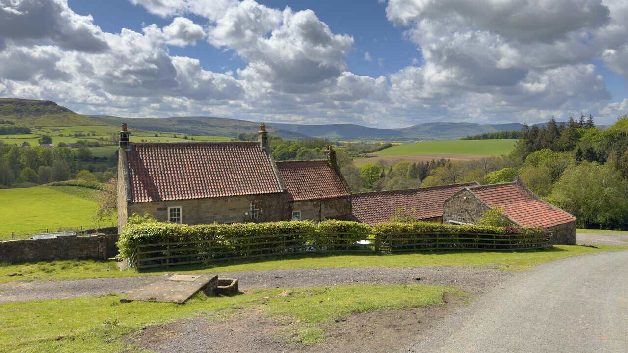 Bankside Farm stands beside a rising tarmac lane on the Great Ayton walk, with honey-coloured stone buildings, red pantile roofs and the Cleveland Hills behind.
