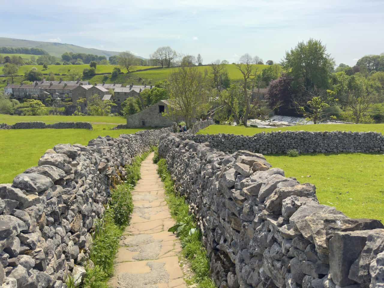 The Sedber Lane footpath leading down to Linton Falls, enclosed by dry stone walls with the Cracoe Fell obelisk visible on the horizon.