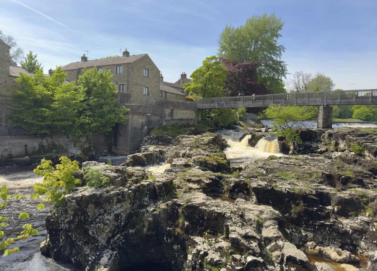 The wooden footbridge at Linton Falls seen from the riverbank, with stone cottages clustered beside the falls and weirs.