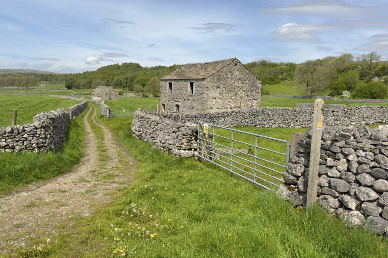 Two large stone barns beside Cove Lane near Grassington, with a wooden public footpath sign pointing the way into Grass Wood.