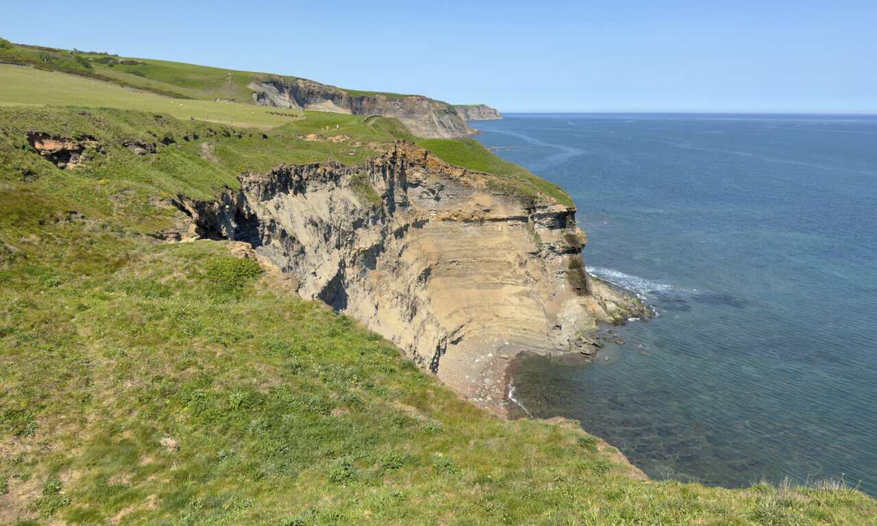 The Cleveland Way looks north towards Craze Naze, with banded cliffs, rocky shore and rolling green clifftops.