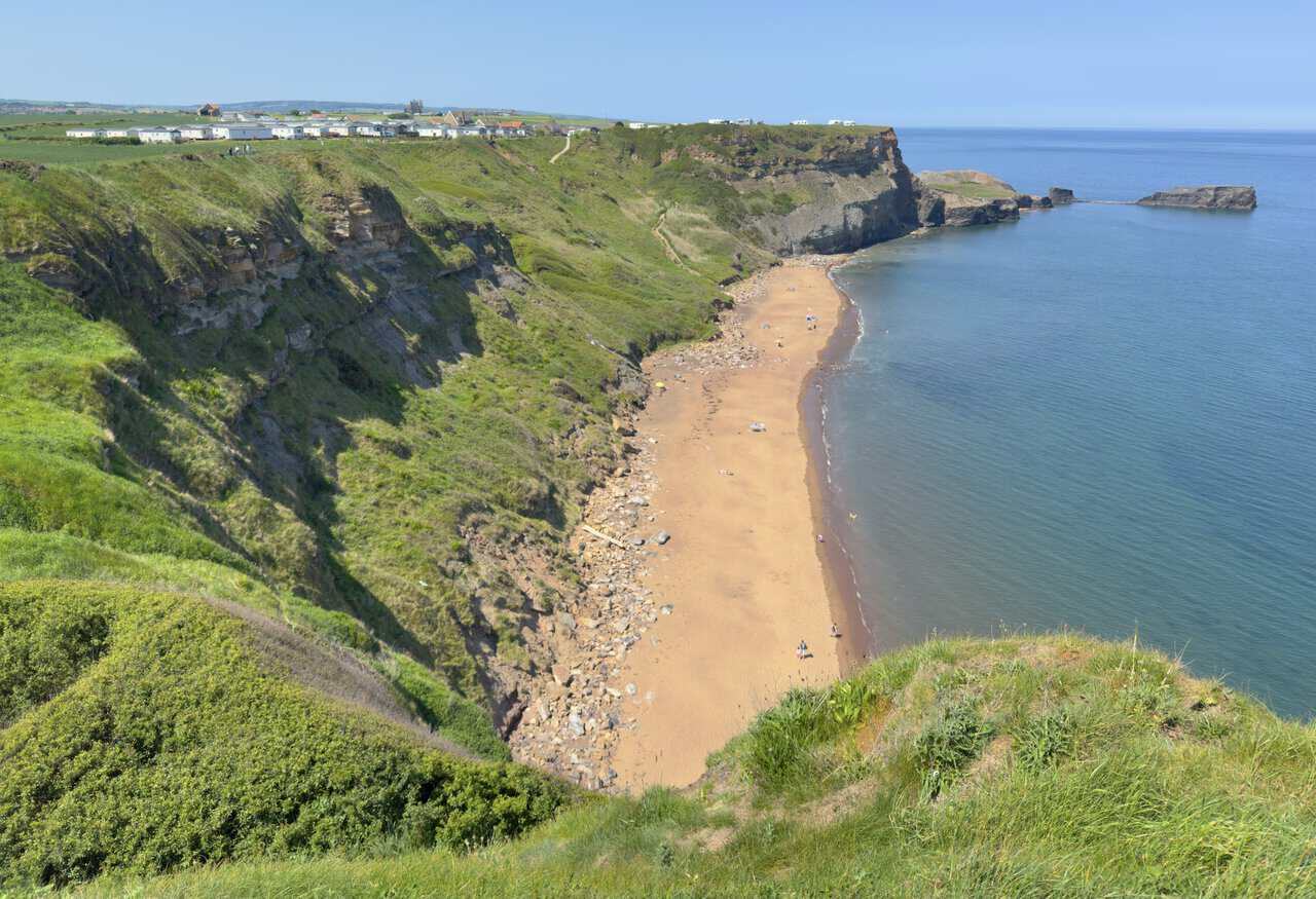 A view down to the golden sands of Saltwick Bay, with Whitby Holiday Park on the clifftop and Whitby Abbey beyond.