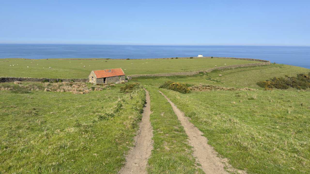 A stone barn with a red pantile roof stands below the Cinder Track, with the sea and a small white clifftop building beyond.