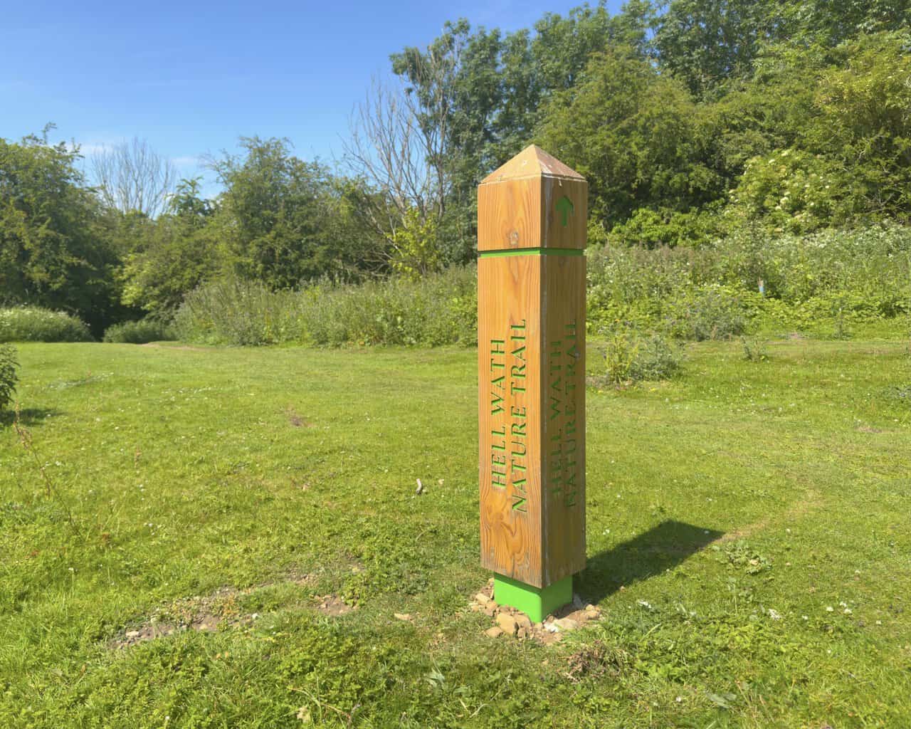 A green waymarker post for the Hell Wath Nature Trail in Ripon, set beside meadow and woodland.