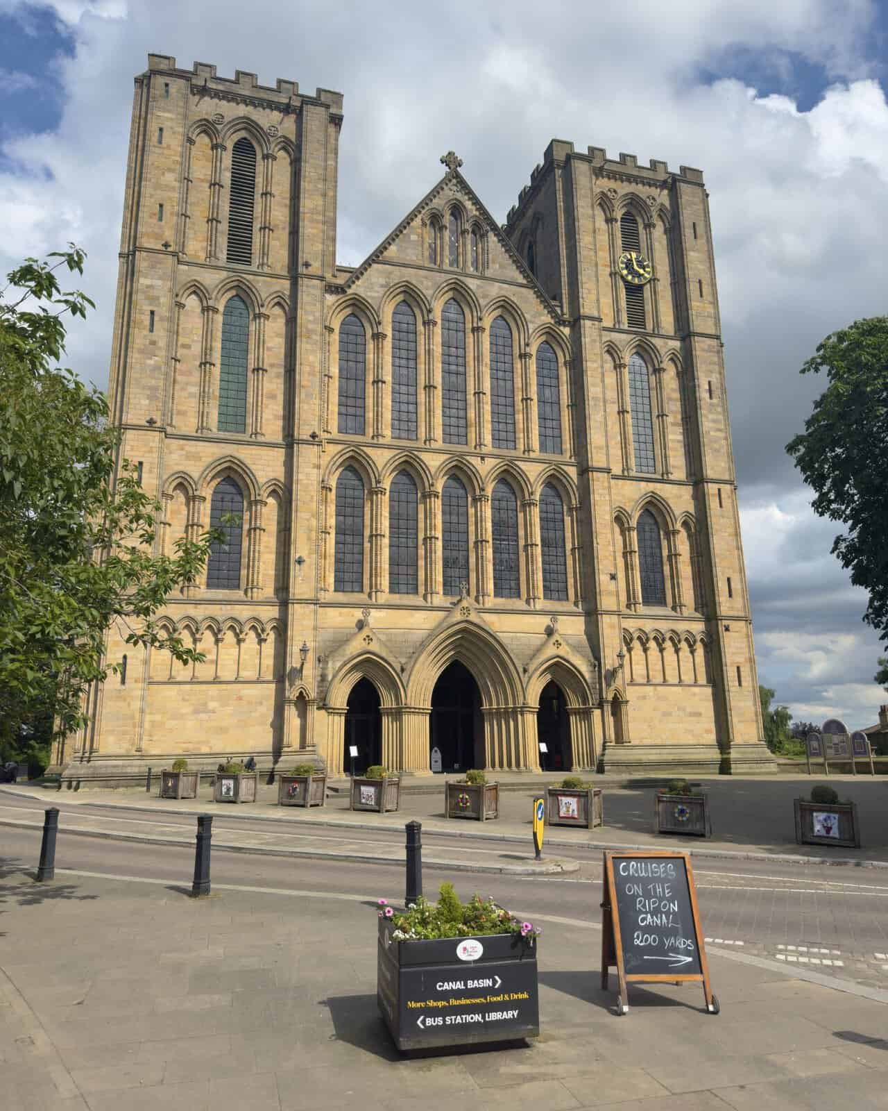 Ripon Cathedral west front at the end of the walk, with twin towers, arched doorways and the gilded clock nearby.
