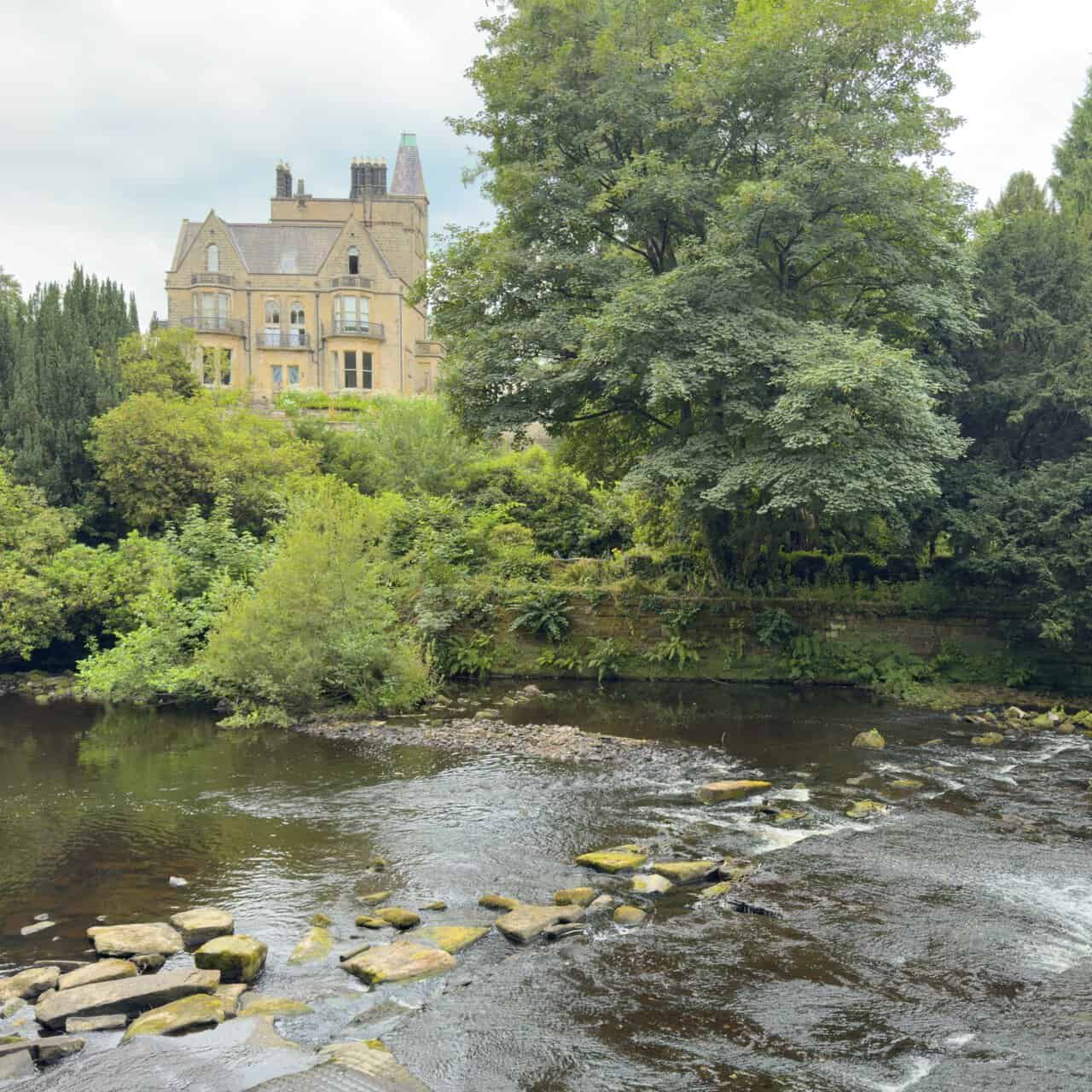 Broad weir across the River Nidd with the turreted Castlestead house among trees above.