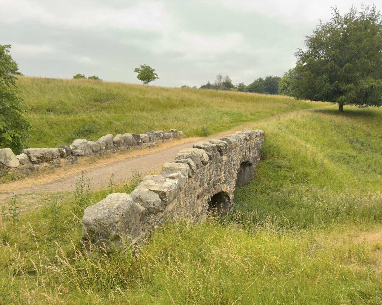 A low stone bridge with parapets of great squared blocks, crossing a dip in the open parkland.