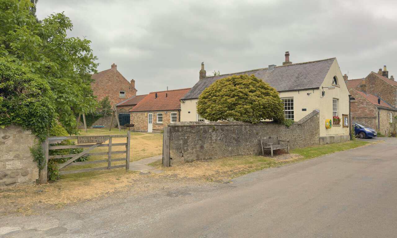 Studley Roger Village Hall, set back behind a high stone wall with a wooden bench beside it.