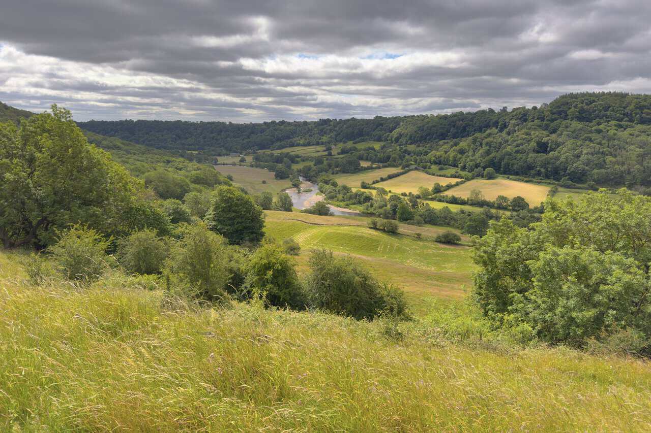 River Swale winding through the valley floor, with green fields, hedgerows and steep wooded slopes on either side.
