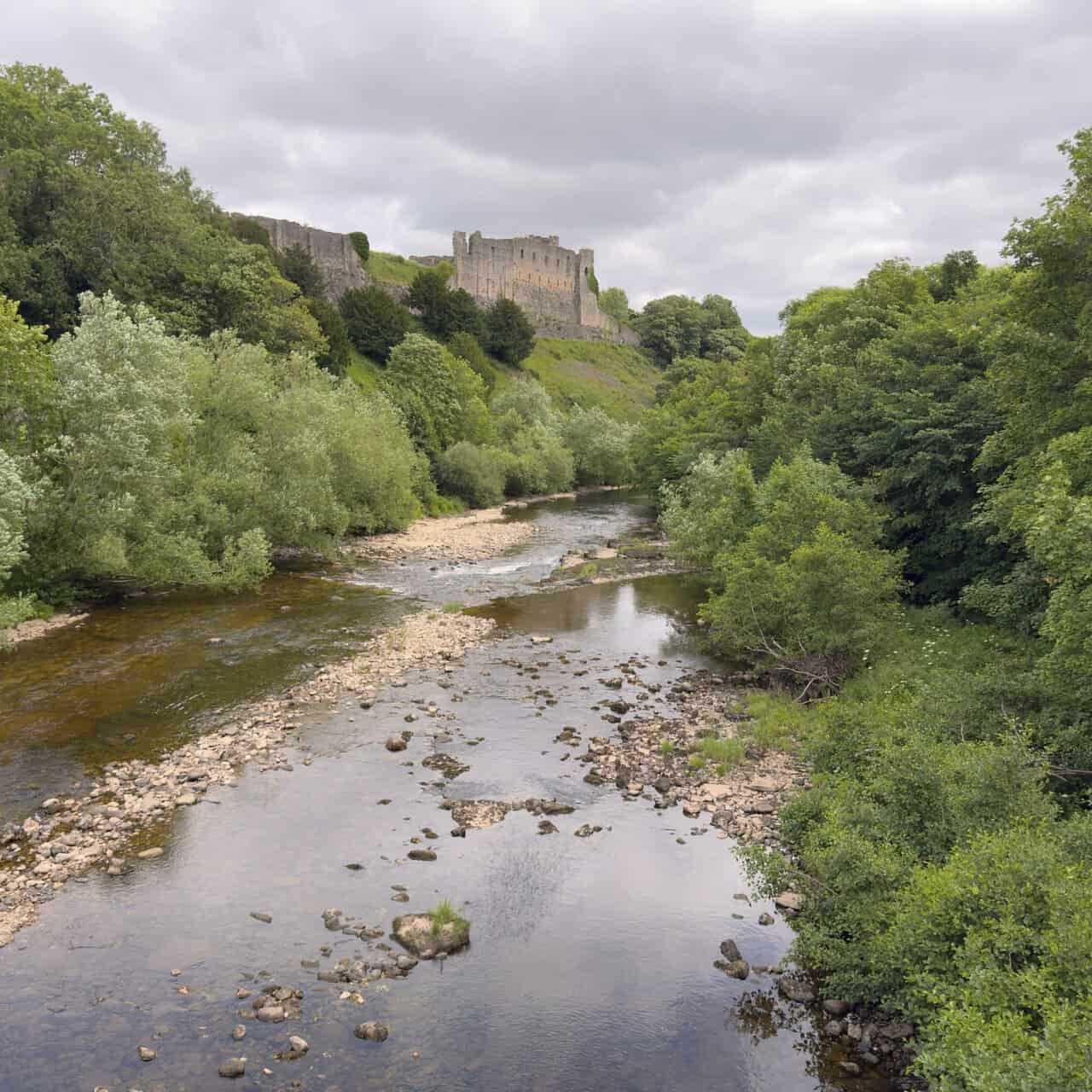 Richmond Castle rising above the River Swale, with shallow water, riverside trees and the castle walls on the cliff.