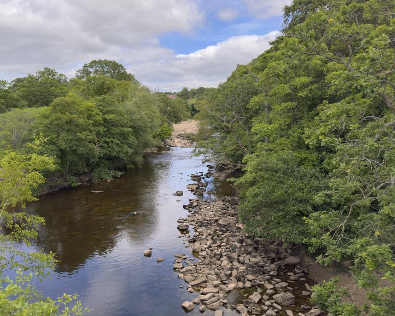 Footbridge over the River Swale on the Richmond walk, with shingle beds, scattered rocks and wooded riverbanks upstream.