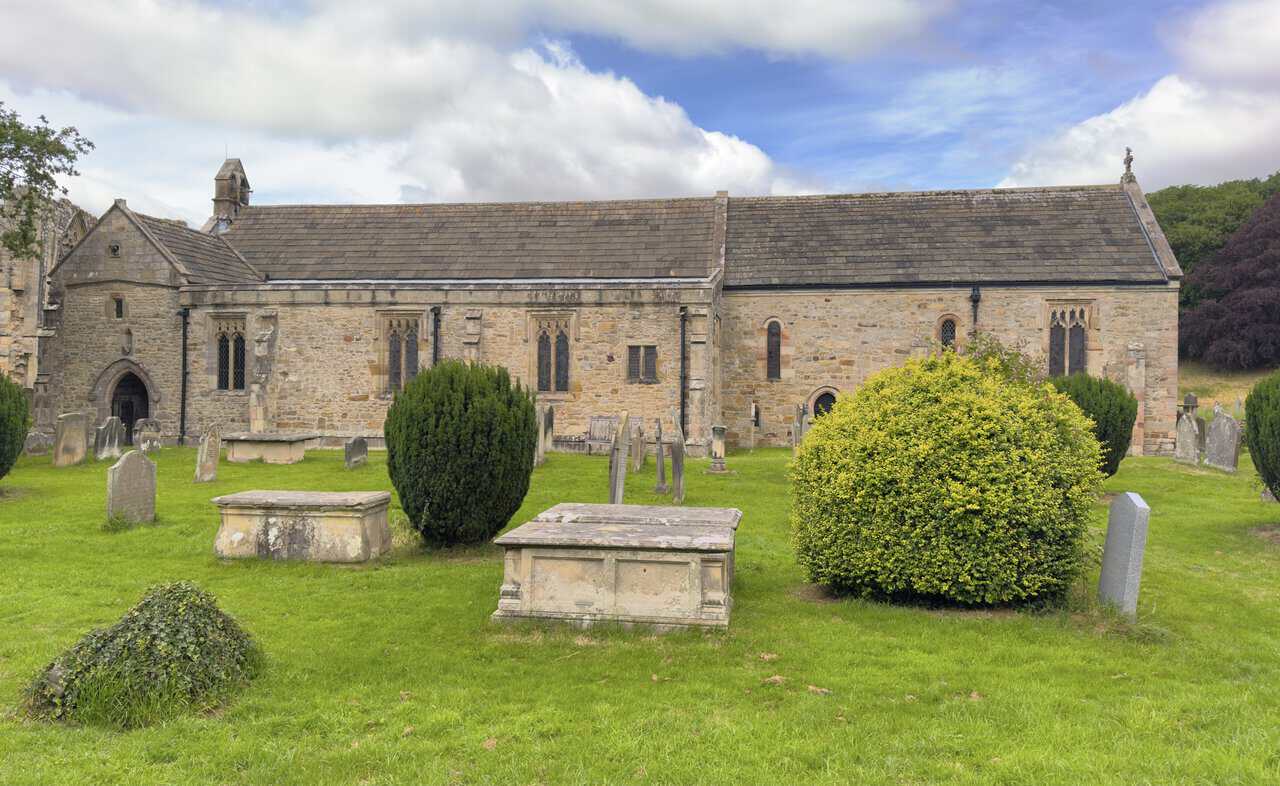 Side view of St Agatha’s Church, with weathered gravestones, stone table tombs and the churchyard beside Easby Abbey.