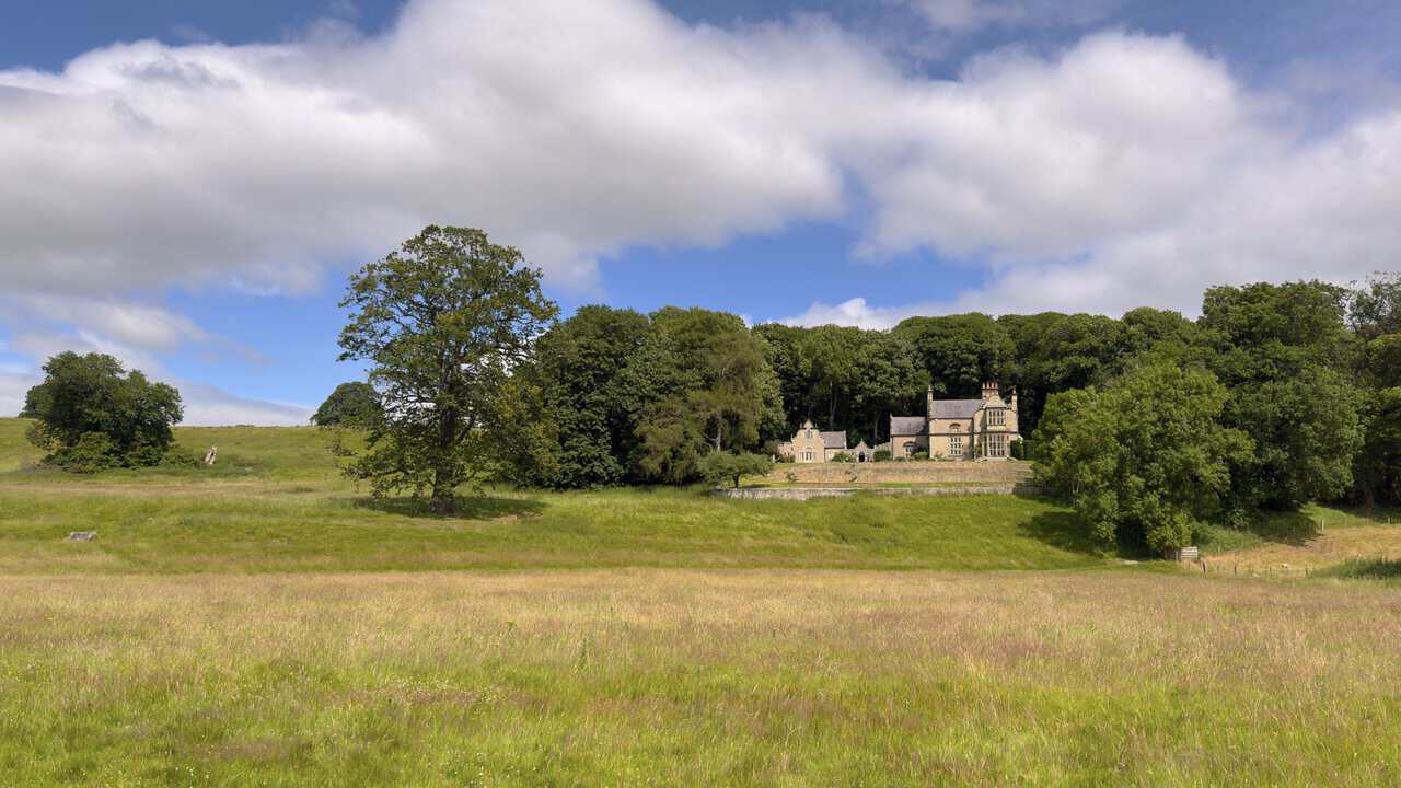 View across a meadow towards a handsome stone house near Abbey Mill, framed by trees and rough grassland.