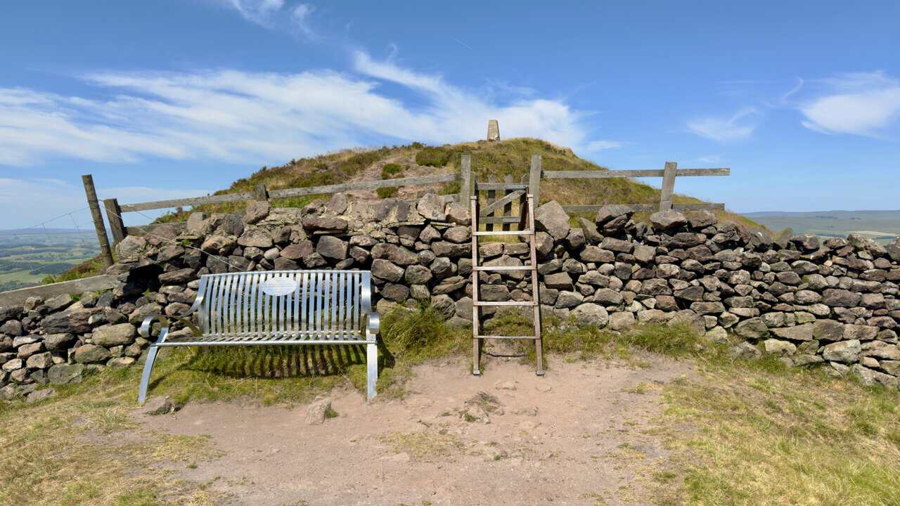 Metal bench and stone stile below the summit of Sharp Haw, with the trig point on the top.