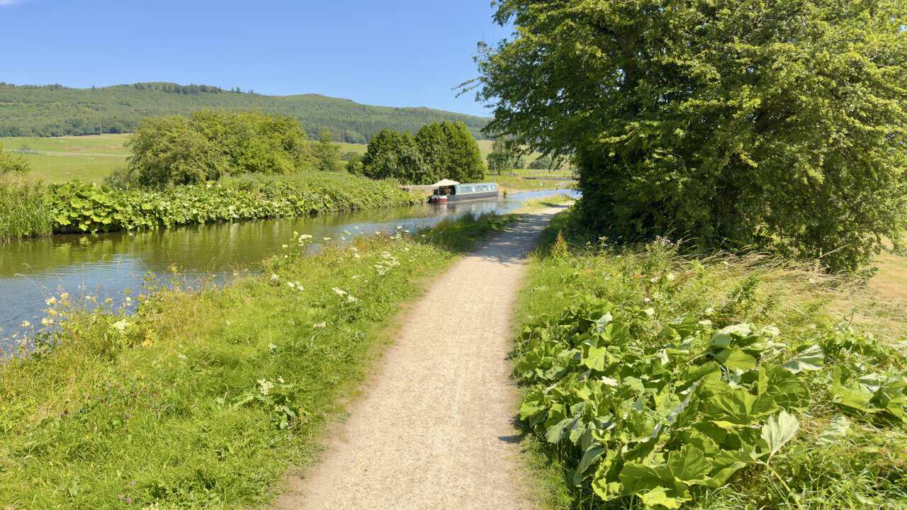 Leeds and Liverpool Canal towpath heading east with a moored narrowboat under the trees.