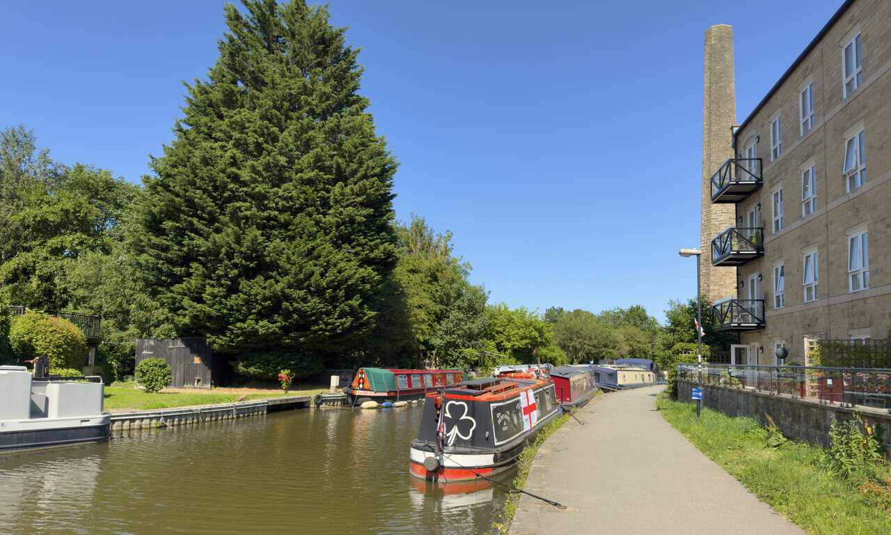 Converted mill flats and a tall stone chimney above narrowboats moored on the canal in Skipton.