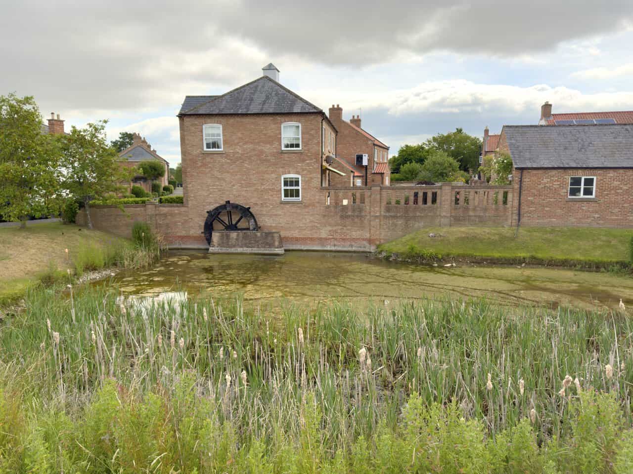 Old waterwheel set into the base of a tall brick house beside a reed-fringed pond in North Stainley.