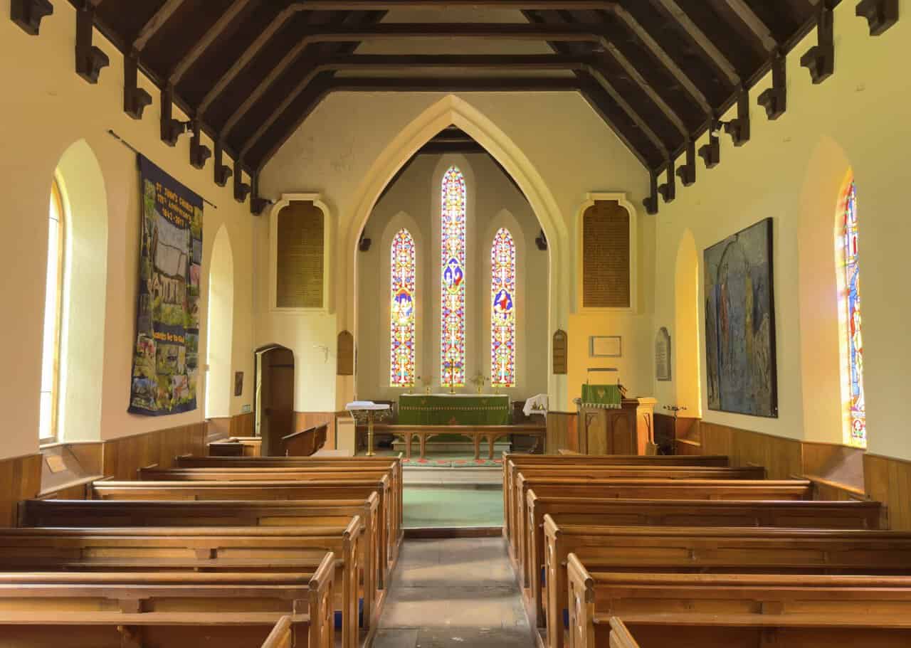 Interior of St John the Evangelist Church with wooden pews, a dark timber roof and three stained-glass windows above the altar.