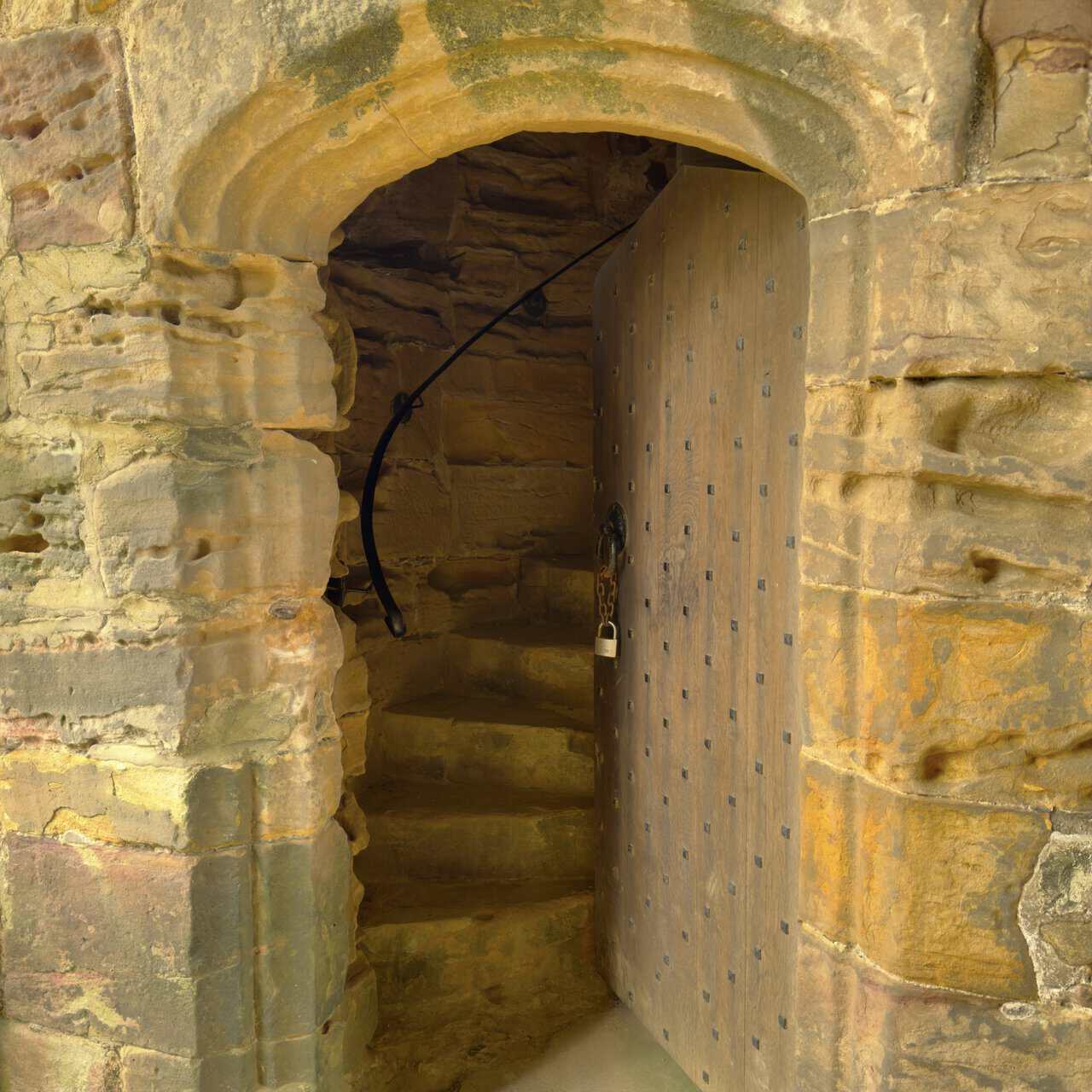 Narrow spiral stone staircase inside Marmion Tower with worn steps, an open studded door and a metal handrail.