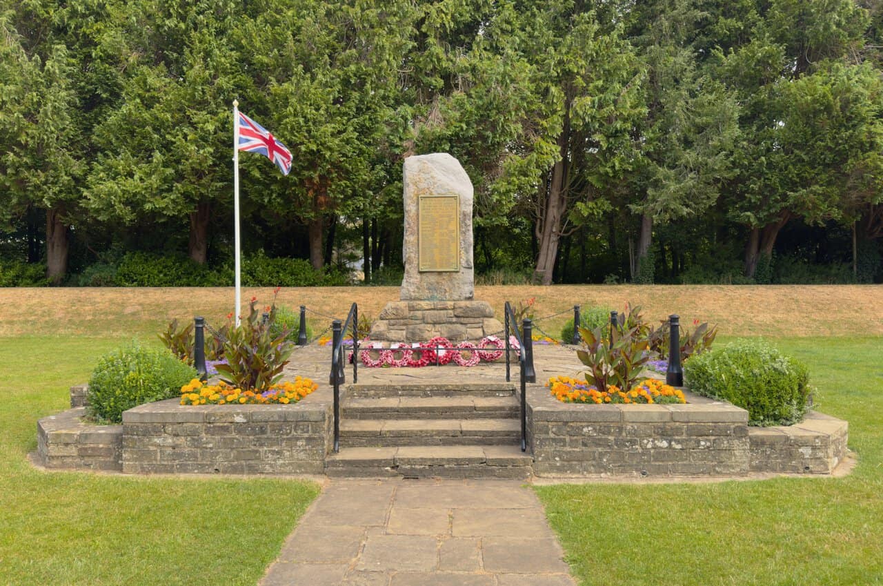 War memorial standing stone at Bewerley Recreation Ground at the start of the Yorke's Folly walk, with a Union flag and colourful flowerbeds.