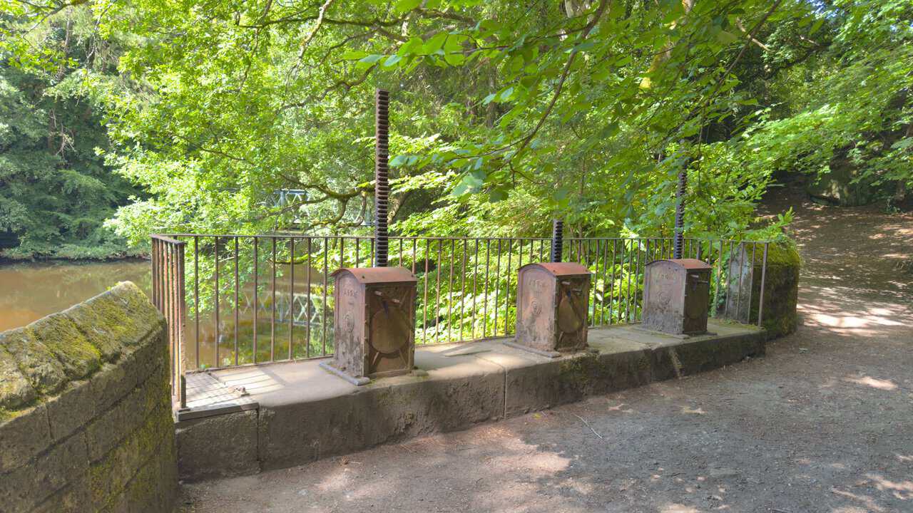 Three old cast-iron sluice mechanisms beside the River Nidd, each with an upright screw thread on a boxed casing.