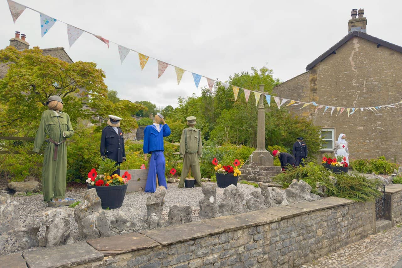 Kettlewell war memorial cross surrounded by scarecrows dressed in wartime service uniforms beneath bunting.