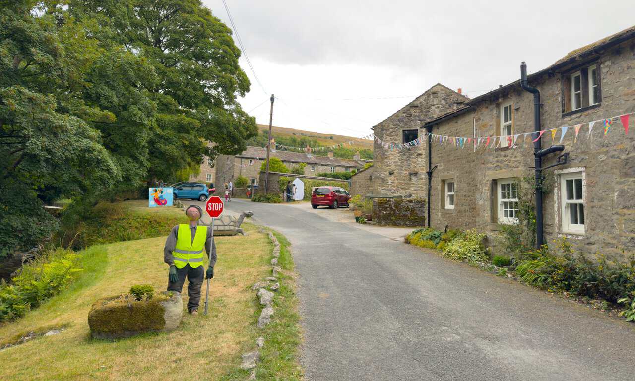 A scarecrow in a high-visibility jacket holding a stop sign beside a village lane on the Great Whernside circular walk.